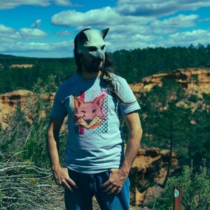 Puede incluir: Una persona con una m&aacute;scara de zorro gris y una camiseta gris con un gr&aacute;fico de zorro. La persona est&aacute; de pie al aire libre con un tel&oacute;n de fondo de &aacute;rboles, colinas y un cielo azul con nubes. La camiseta presenta un dise&ntilde;o de zorro rosa y naranja.