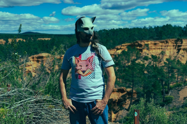 Puede incluir: Una persona con una m&aacute;scara de zorro gris y una camiseta gris con un gr&aacute;fico de zorro. La persona est&aacute; de pie al aire libre con un tel&oacute;n de fondo de &aacute;rboles, colinas y un cielo azul con nubes. La camiseta presenta un dise&ntilde;o de zorro rosa y naranja.