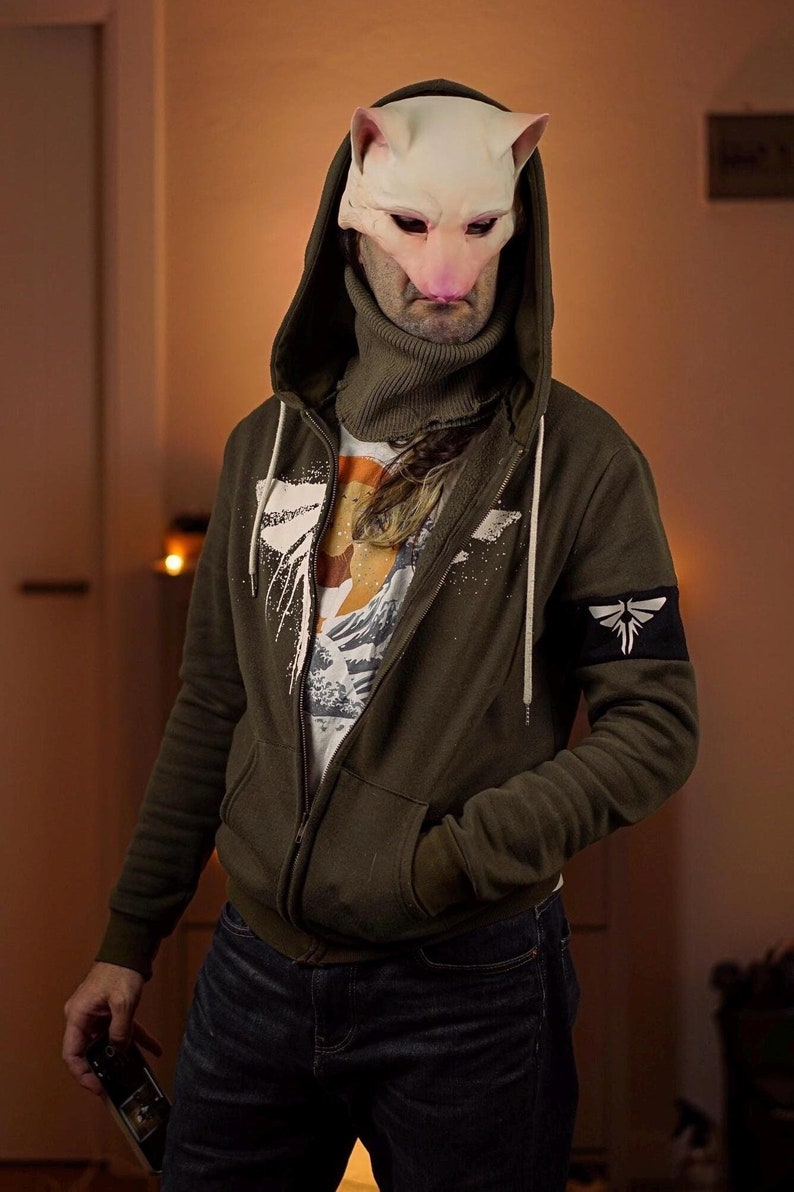 Op de afbeelding: Een persoon draagt ​​een groene hoodie met een witte grafische print en een witte rits. De hoodie heeft een zwarte patch op de linkermouw met een wit libellenontwerp. De persoon draagt ​​ook een blauwe jeans en een witte turtleneck. De persoon draagt ​​een wit kattenmasker met roze oren en neus.