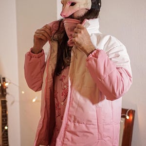 Op de afbeelding: Een roze en witte pufferjas met een ritssluiting. De jas heeft een capuchon en een witte kraag. De persoon die de jas draagt, draagt eronder een shirt met bloemenprint.