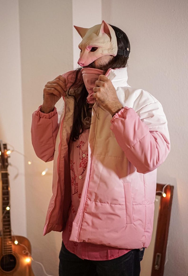 Op de afbeelding: Een roze en witte pufferjas met een ritssluiting. De jas heeft een capuchon en een witte kraag. De persoon die de jas draagt, draagt eronder een shirt met bloemenprint.
