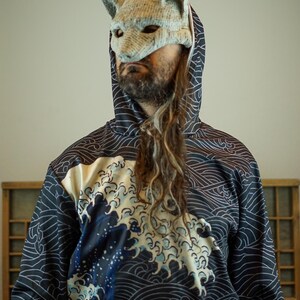 Op de afbeelding: Een persoon draagt een marineblauwe hoodie met een wit golfpatroon en een grote golfafbeelding in tinten blauw en wit. De persoon draagt ook een vossenmasker van licht materiaal.
