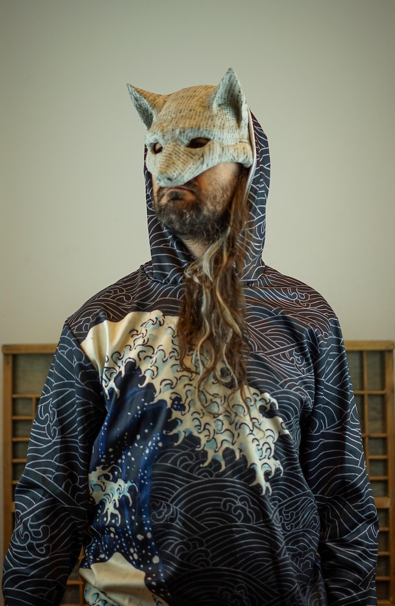 Op de afbeelding: Een persoon draagt een marineblauwe hoodie met een wit golfpatroon en een grote golfafbeelding in tinten blauw en wit. De persoon draagt ook een vossenmasker van licht materiaal.