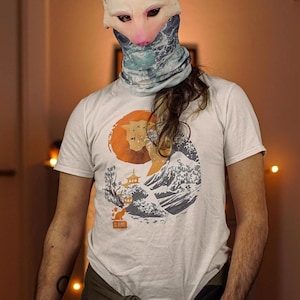 Op de afbeelding: Een wit T-shirt met een afbeelding van een kat voor een golf en een Japanse pagode. De kat is oranje en de golf is blauw en wit. De achtergrond is een rode cirkel.