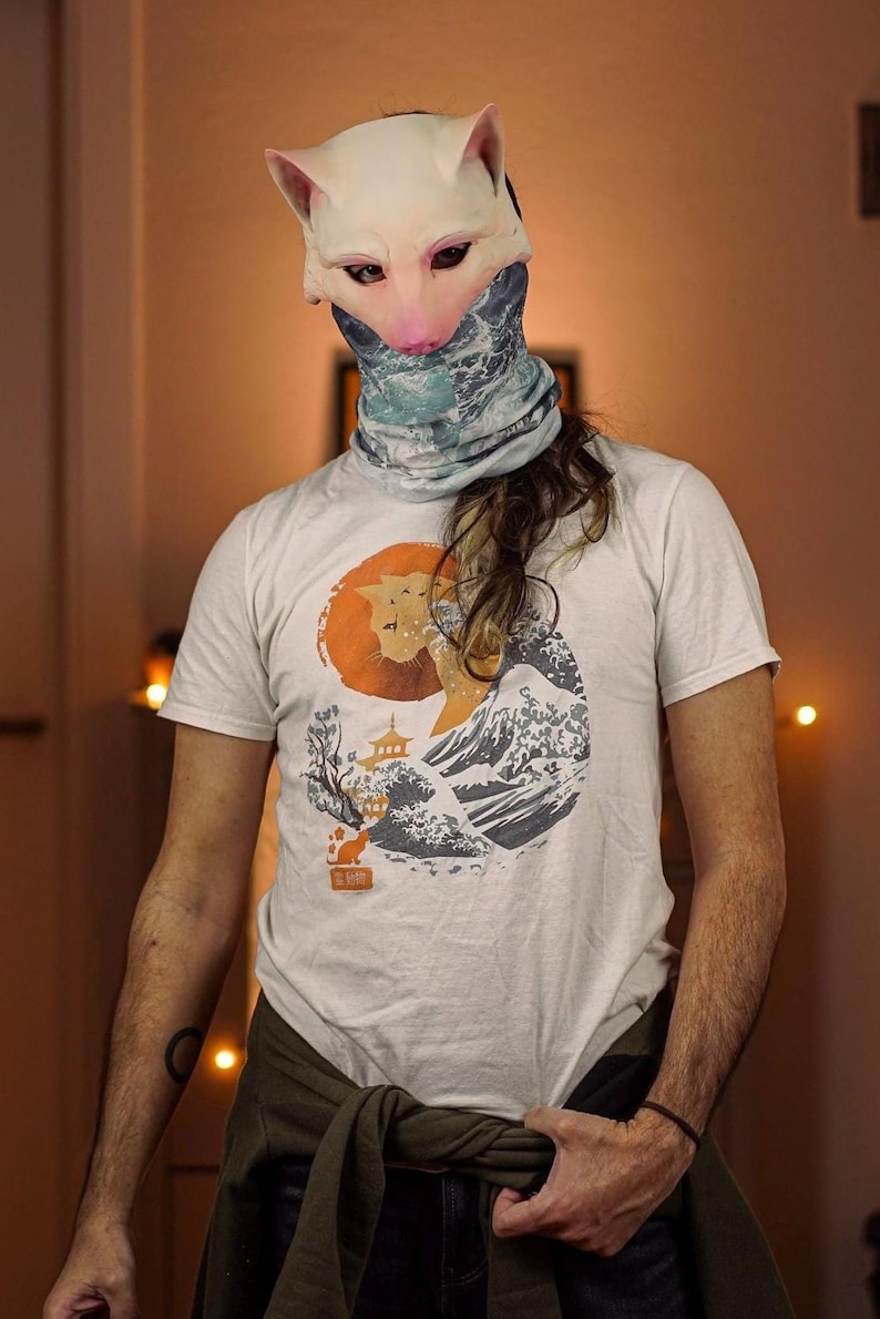 Op de afbeelding: Een wit T-shirt met een afbeelding van een kat voor een golf en een Japanse pagode. De kat is oranje en de golf is blauw en wit. De achtergrond is een rode cirkel.