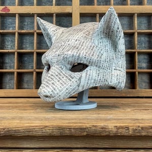 Op de afbeelding: Een handgemaakt kattenmasker van krantenpapier, met puntige oren en oogopeningen. Het masker staat op een grijze standaard, waarbij de textuur van het papier en de gedrukte tekst zichtbaar zijn. De achtergrond is van hout.