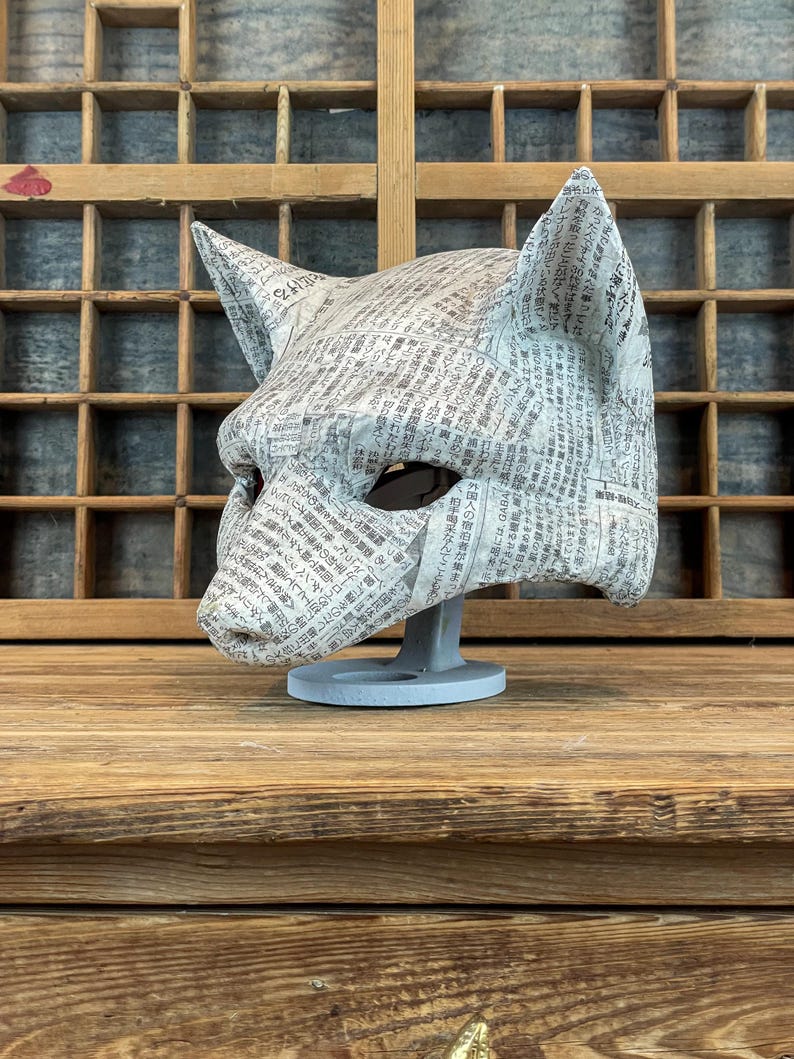Op de afbeelding: Een handgemaakt kattenmasker van krantenpapier, met puntige oren en oogopeningen. Het masker staat op een grijze standaard, waarbij de textuur van het papier en de gedrukte tekst zichtbaar zijn. De achtergrond is van hout.
