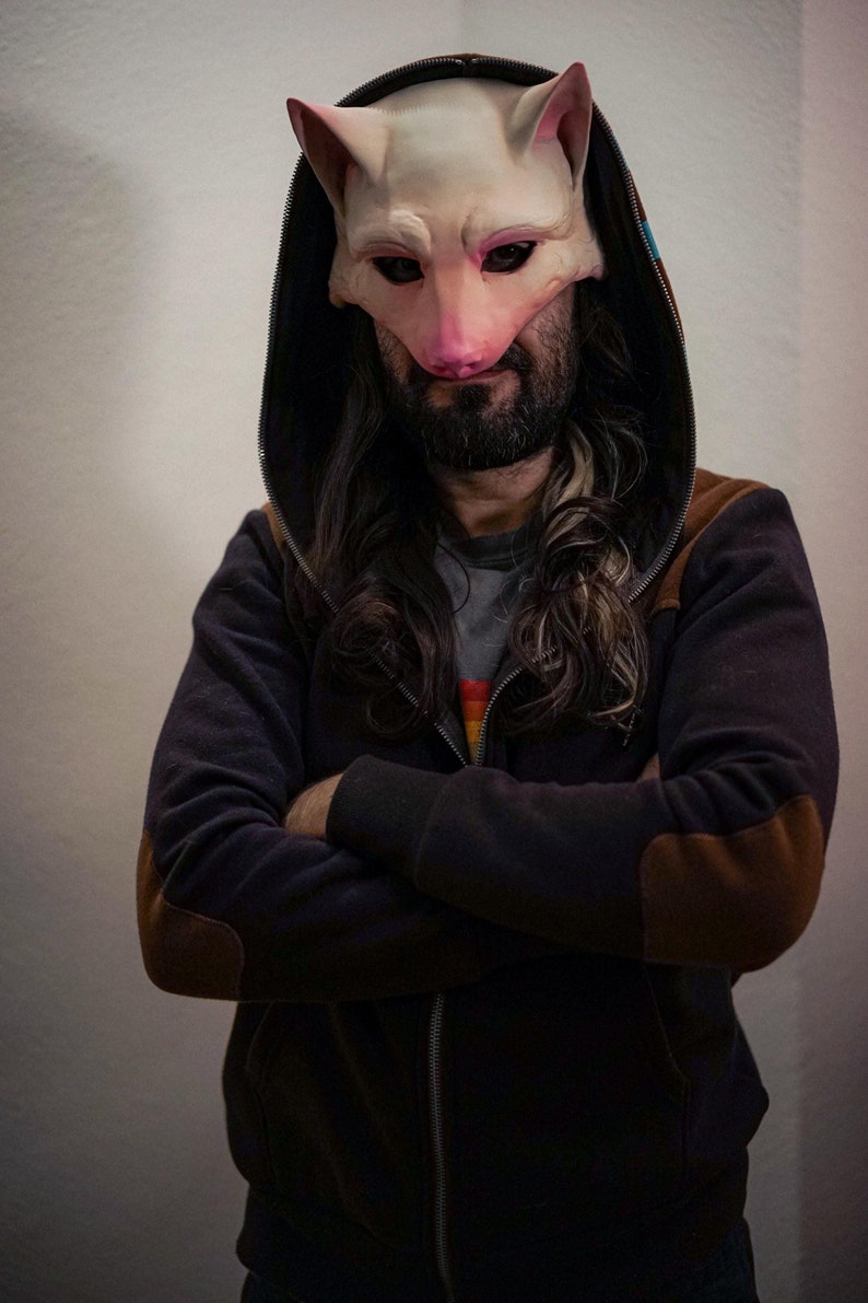 Puede incluir: Una persona con una sudadera con capucha negra con parches de cuero marrón en los codos y una máscara de gato blanca. La máscara tiene detalles rosas alrededor de los ojos y la nariz.