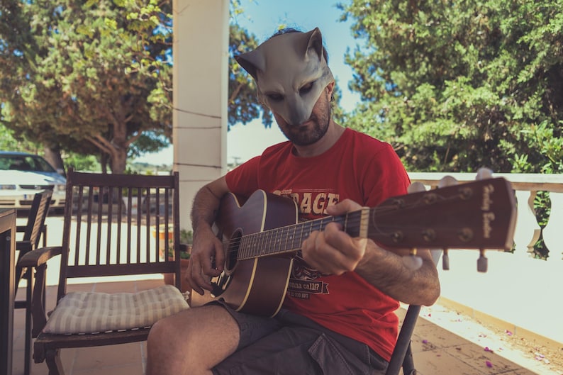 Puede incluir: Una persona con una máscara de lobo gris toca una guitarra acústica mientras está sentada en un porche. La guitarra es negra con una tapa de madera marrón y tiene una golpeador blanco. La persona lleva una camiseta roja con la palabra "Stage" impresa en ella.