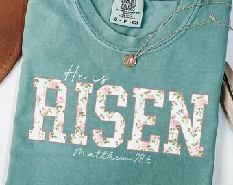 Puede incluir: Una camiseta azul turquesa con un estampado floral blanco que dice "He is RISEN" con la referencia bíblica "Matthew 28:6" debajo.