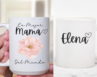 May include: Two white ceramic mugs with black text and a pink flower design. One mug says "La Mejor Mamá" with a heart and the other mug says "Elena" with a heart.