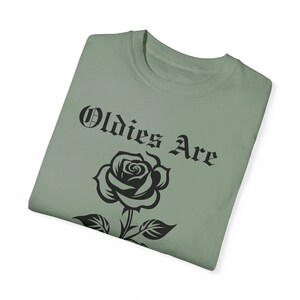 May include: A sage green t-shirt featuring a black rose and the text "Oldies Are" in a gothic font.
