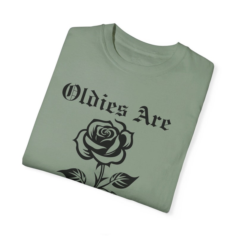 May include: A sage green t-shirt featuring a black rose and the text "Oldies Are" in a gothic font.
