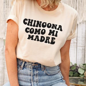 May include: A white t-shirt with black text that reads "Chingona como mi madre" in a retro font.