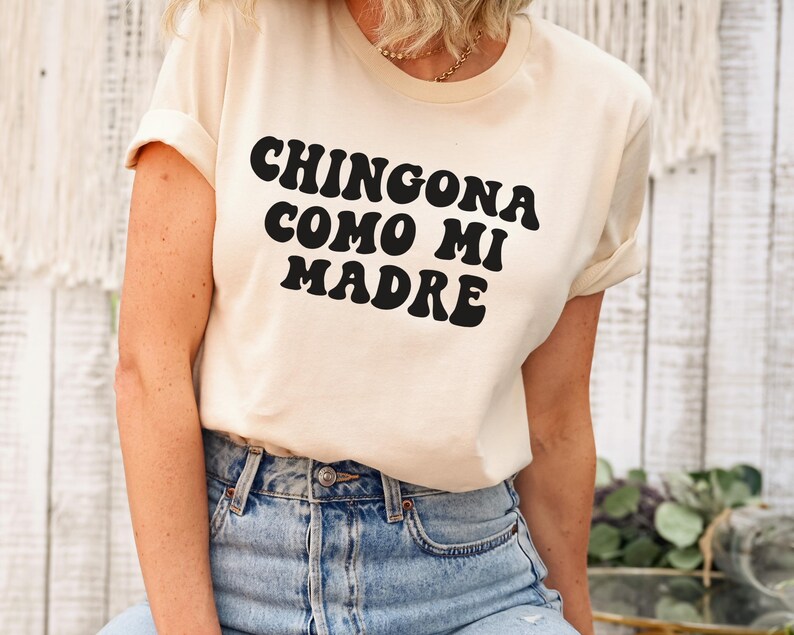 May include: A white t-shirt with black text that reads "Chingona como mi madre" in a retro font.
