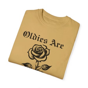 May include: A tan t-shirt featuring a black rose and the text "Oldies Are" in a black gothic font.