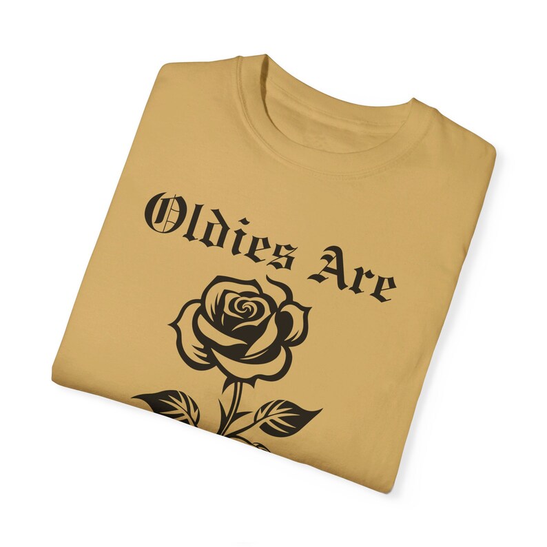 May include: A tan t-shirt featuring a black rose and the text "Oldies Are" in a black gothic font.