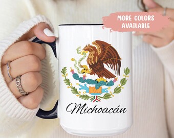 May include: White ceramic mug with a black handle and rim, featuring the coat of arms of Michoacán, Mexico. The design includes a brown eagle, a snake, and the text "Michoacán". The text "MORE COLORS AVAILABLE" is also visible.