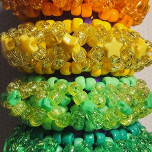 K&ouml;nnte beinhalten: Vier bunte Perlenarmb&auml;nder, die &uuml;bereinander gestapelt sind. Das oberste Armband ist orange, gefolgt von gelb, gr&uuml;n und blaugr&uuml;n. Jedes Armband hat ein anderes Design mit sternf&ouml;rmigen Perlen.