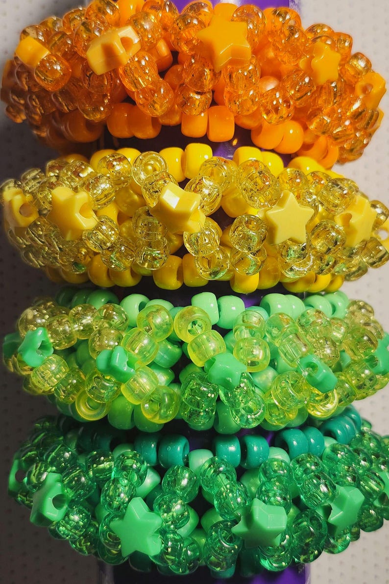 K&ouml;nnte beinhalten: Vier bunte Perlenarmb&auml;nder, die &uuml;bereinander gestapelt sind. Das oberste Armband ist orange, gefolgt von gelb, gr&uuml;n und blaugr&uuml;n. Jedes Armband hat ein anderes Design mit sternf&ouml;rmigen Perlen.