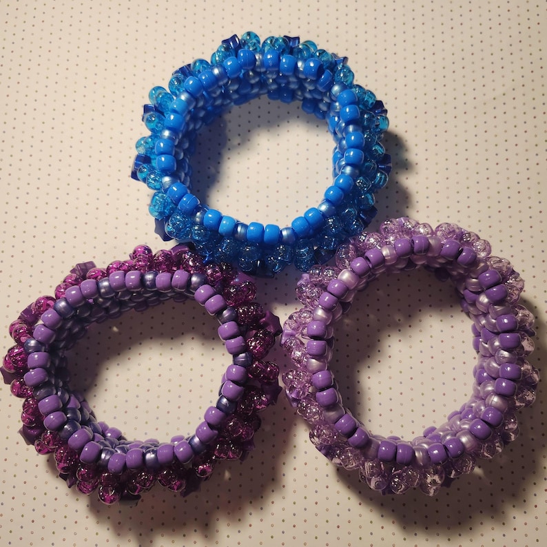 K&ouml;nnte beinhalten: Drei Armb&auml;nder aus Perlen in Blau- und Lilat&ouml;nen. Die Armb&auml;nder bestehen aus kleinen, runden Perlen mit einem mehrschichtigen Design. Das blaue Armband befindet sich oben, darunter zwei lila Armb&auml;nder. Die Armb&auml;nder sind wahrscheinlich handgefertigt.