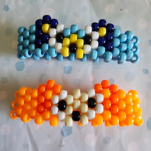 Puede incluir: Dos pulseras de cuentas. La pulsera superior es azul claro, azul oscuro, amarillo, negro y blanco. La pulsera inferior es naranja, blanca y negra. Las pulseras están hechas de pequeñas cuentas redondas.