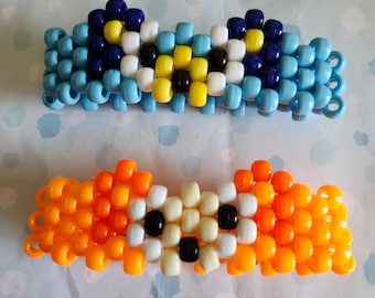 Könnte beinhalten: Zwei Armbänder aus Perlen. Das obere Armband ist hellblau, dunkelblau, gelb, schwarz und weiß. Das untere Armband ist orange, weiß und schwarz. Die Armbänder bestehen aus kleinen, runden Perlen.