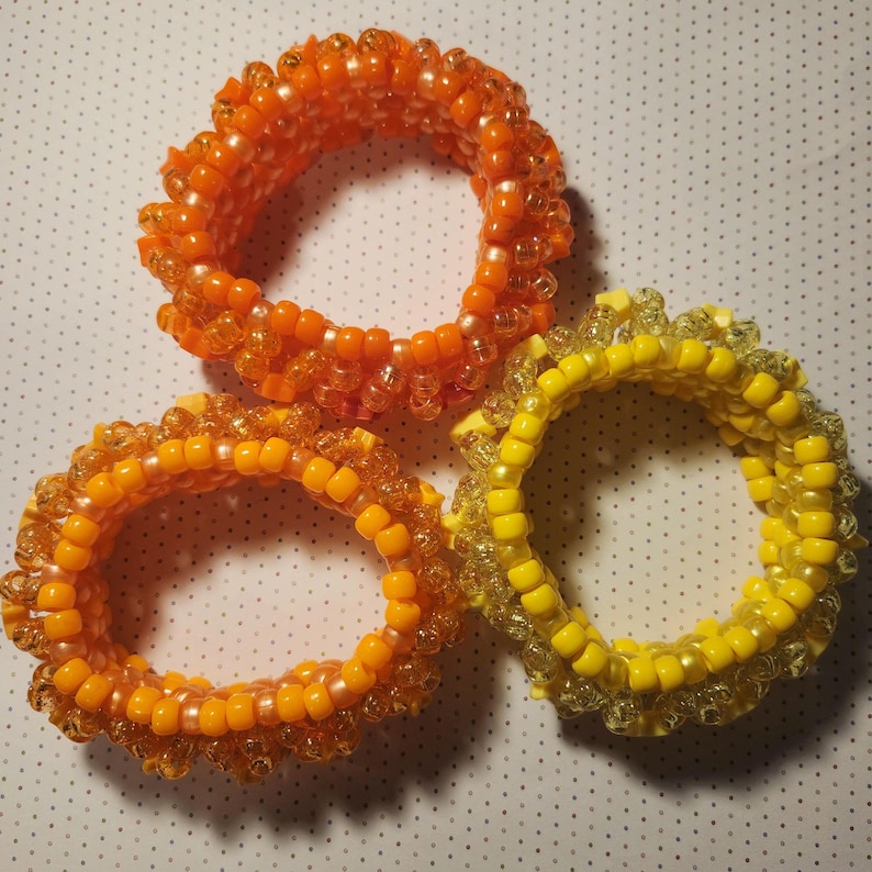 K&ouml;nnte beinhalten: Drei Armb&auml;nder aus Perlen in Orange- und Gelbt&ouml;nen. Die Armb&auml;nder bestehen aus kleinen, runden Perlen, und die Farben reichen von einem hellen, sonnigen Gelb bis zu einem tiefen, gebrannten Orange. Die Armb&auml;nder sind kreisf&ouml;rmig angeordnet.