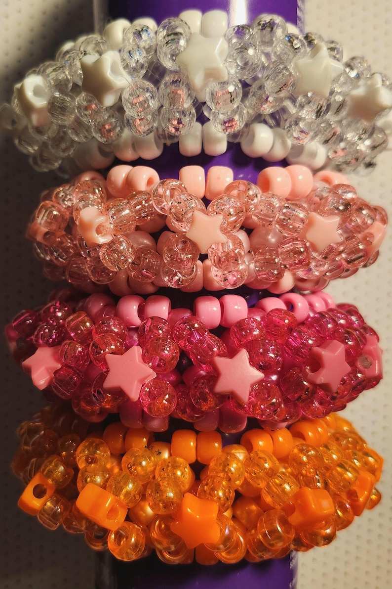 K&ouml;nnte beinhalten: Gestapelte, farbenfrohe Armb&auml;nder aus Perlen in den Farbt&ouml;nen Klar, Rosa und Orange. Die Armb&auml;nder haben sternf&ouml;rmige und quadratische Perlen. Die Armb&auml;nder sind auf einem violetten zylindrischen Objekt gestapelt.