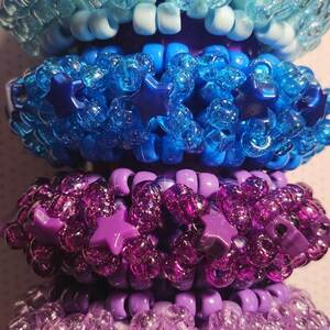 K&ouml;nnte beinhalten: Vier farbenfrohe Armb&auml;nder aus Perlen in Blau- und Lilat&ouml;nen. Das oberste Armband ist hellblau mit sternf&ouml;rmigen Perlen. Das zweite Armband ist dunkelblau mit Sternakzenten. Das dritte und vierte Armband sind lila mit sternf&ouml;rmigen Perlen.