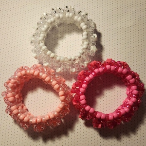 K&ouml;nnte beinhalten: Drei Armb&auml;nder aus Perlen in Rosa- und Wei&szlig;t&ouml;nen. Die Armb&auml;nder bestehen aus kleinen, runden Perlen und haben einen ger&uuml;schten Rand. Ein Armband ist wei&szlig;, eines hellrosa und das andere dunkler rosa.