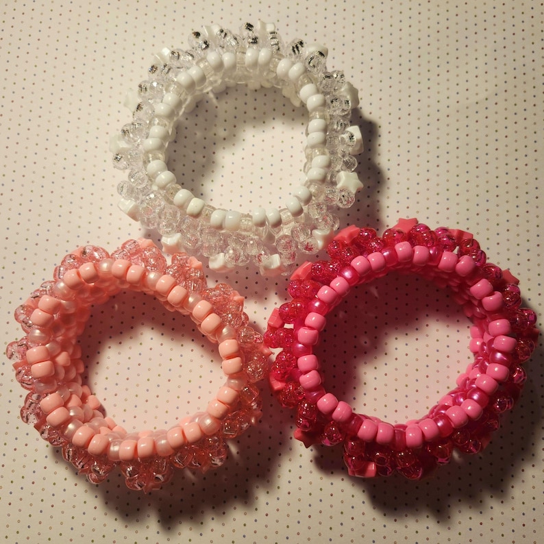 K&ouml;nnte beinhalten: Drei Armb&auml;nder aus Perlen in Rosa- und Wei&szlig;t&ouml;nen. Die Armb&auml;nder bestehen aus kleinen, runden Perlen und haben einen ger&uuml;schten Rand. Ein Armband ist wei&szlig;, eines hellrosa und das andere dunkler rosa.
