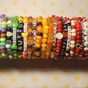 May include: A stack of colourful beaded bracelets with words such as "Sour", "Caution", "Your", "Mom", "Circus", "Lovely", "Noel", "Grubel", "Charlie", "Mush", "Ram", and "Elise" spelled out.