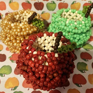 Può includere: Tre contenitori a forma di mela realizzati con piccole perline di plastica. Uno è rosso, uno verde chiaro e uno dorato, ognuno con un interno bianco, un gambo marrone e un piccolo dettaglio a foglia verde sul bordo superiore della struttura.