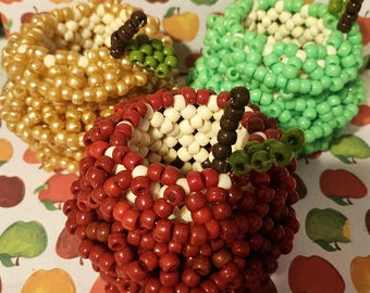 Può includere: Tre contenitori a forma di mela realizzati con piccole perline di plastica. Uno è rosso, uno verde chiaro e uno dorato, ognuno con un interno bianco, un gambo marrone e un piccolo dettaglio a foglia verde sul bordo superiore della struttura.