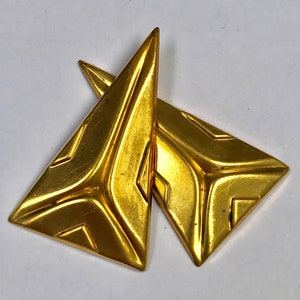 May include: A pair of gold-tone geometric earrings with a triangular shape. The earrings have a textured surface and a modern design.