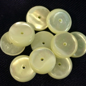 May include: A collection of pale yellow, round beads with a pearlescent sheen. Each bead has a central hole, and they vary slightly in size. The beads are scattered on a dark surface, creating a contrast that highlights their color and texture.