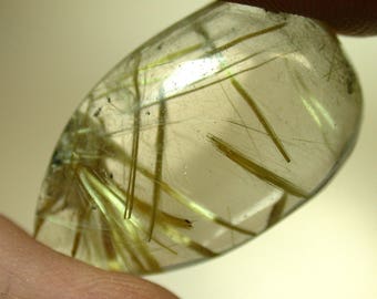 Op de afbeelding: Een heldere kwartscabochon met gouden rutile-insluitsels. De rutile-insluitsels zijn zichtbaar in de hele steen, waardoor een glinsterend effect ontstaat.