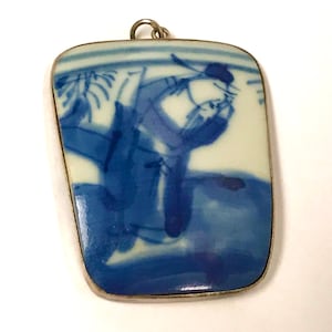 May include: A blue and white porcelain pendant with a silver frame. The pendant features a hand-painted design of a person in traditional Chinese clothing.