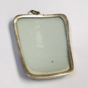May include: A silver pendant with a rectangular frame and a white porcelain insert. The pendant has a loop at the top for attaching a chain.