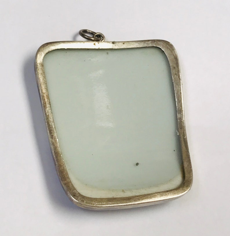 May include: A silver pendant with a rectangular frame and a white porcelain insert. The pendant has a loop at the top for attaching a chain.