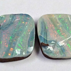 Könnte beinhalten: Zwei schillernde Opal-Edelsteine in hellblau und grün. Die Opale sind in einer dunkelbraunen Fassung gefasst.