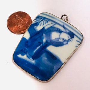 May include: A silver pendant with a rectangular blue and white porcelain tile featuring a Chinese scene of a person riding a horse. The tile is surrounded by a silver frame.