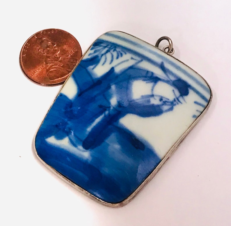 May include: A silver pendant with a rectangular blue and white porcelain tile featuring a Chinese scene of a person riding a horse. The tile is surrounded by a silver frame.