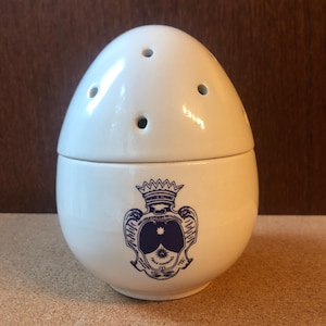 Może przedstawiać: Biały ceramiczny pojemnik w kształcie jaja z pokrywką z czterema małymi otworami. Dolna część przedstawia niebieski wzór herbu. Ten element dekoracyjny może być solniczką lub podobnym przedmiotem kuchennym.