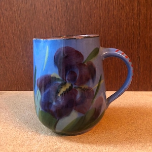 Puede incluir: Una taza de cerámica con un fondo azul y un gran diseño de flor de iris púrpura. El asa es azul con puntos rojos. La taza tiene un borde dorado y hojas verdes. La taza está sobre una superficie beige.