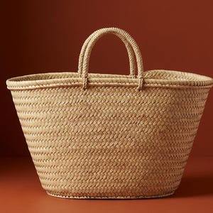 Peut inclure: Un sac fourre-tout en paille tressée de couleur naturelle, avec deux anses arrondies. Le sac présente un motif de tissage en forme de panier et un large dessus ouvert. Le sac est de couleur beige et posé sur un fond brun rougeâtre.