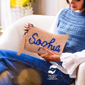 May include: A rectangular burlap clutch with the name "Sophia" embroidered in blue script. The clutch has a matching wrist strap and a gold-colored zipper. The image also shows the logo "Instant Craft Cadeaux".