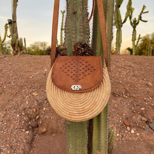 Peut inclure: Un sac bandoulière rond en paille tressée avec un rabat et une bandoulière en cuir marron. Le rabat présente un motif gaufré et un fermoir argenté. Le sac est suspendu devant un cactus.