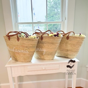 Peut inclure: Trois grands sacs cabas en paille naturelle tissée avec des anses en cuir brun. Les sacs sont remplis de fleurs et portent des inscriptions blanches sur le devant. Les sacs sont posés sur une table blanche.